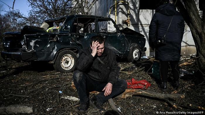 Чоловік сидить поруч із зруйнованим автомобілем та житловим будинком після ракетного удару та атак бойових вертольотів у місті Чугуїв у Харківській області